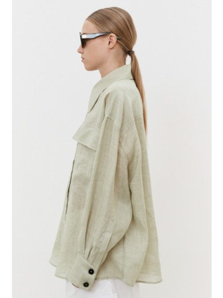 A person with brown hair in a low ponytail faces away, wearing an oversized MEIMEIJ - LINEN SHIRT. Against a plain white background, breathable linen pants are visible at the bottom.