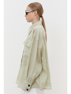 A person with brown hair in a low ponytail faces away, wearing an oversized MEIMEIJ - LINEN SHIRT. Against a plain white background, breathable linen pants are visible at the bottom.