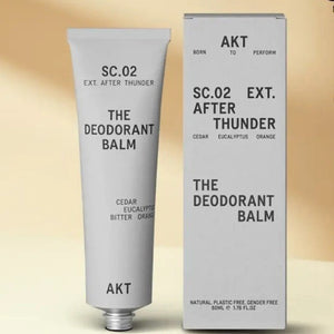 A tube and box of AKT LONDON AKT DEODORANT BALM AFTER THUNDER, featuring cedar, eucalyptus, and orange, provide all-day protection. The minimalist packaging highlights its aluminum-free formula on a beige background.