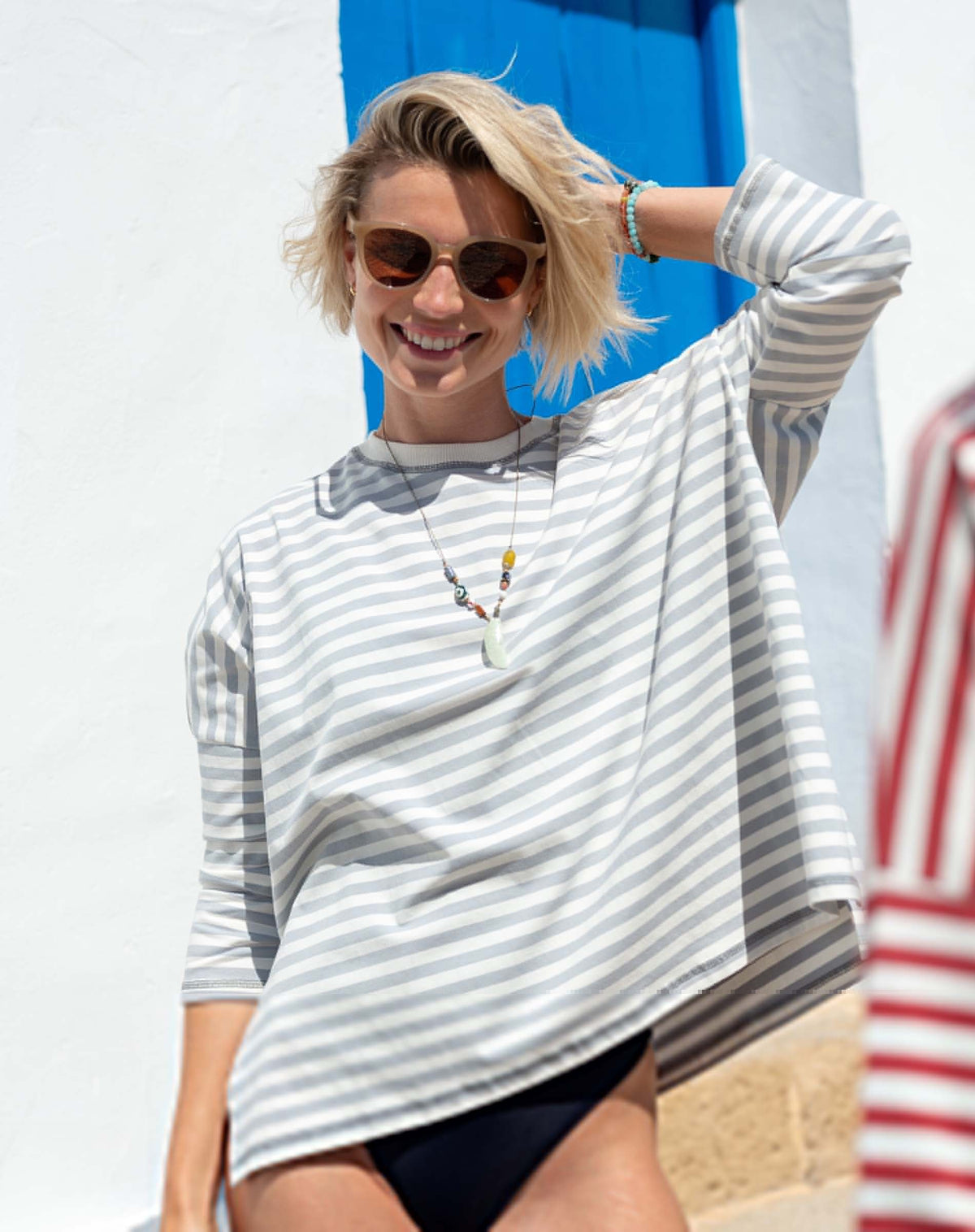 A smiling person with short blonde hair and sunglasses is wearing the MERSEA - CATALINA JERSEY TEE IN SLATE BLUE by MERSEA, along with a necklace and bracelets, standing outdoors in front of a blue door and white wall.