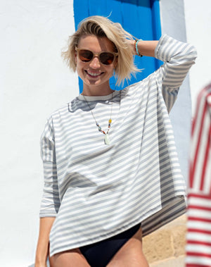A smiling person with short blonde hair and sunglasses is wearing the MERSEA - CATALINA JERSEY TEE IN SLATE BLUE by MERSEA, along with a necklace and bracelets, standing outdoors in front of a blue door and white wall.