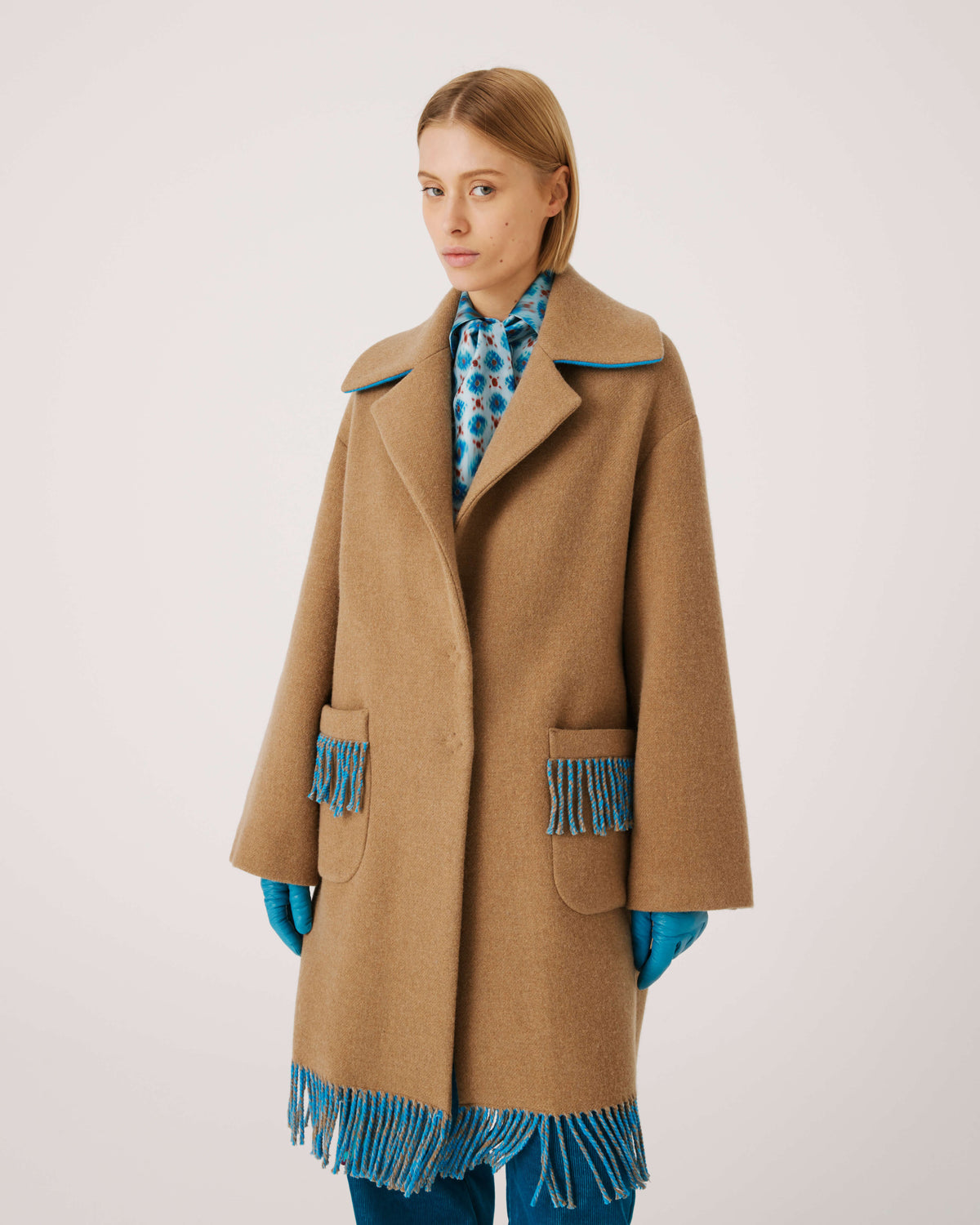 A person is wearing the SFIZIO - TWO TONED COAT WITH FRINGE by SFIZIO, featuring blue and brown fringe patch pockets and hem. They are also donning blue gloves and blue pants. The image is cropped to show from the shoulders down against a plain, off-white background.