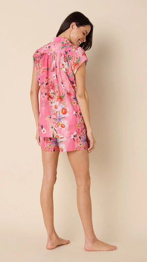 A woman smiles barefoot against a beige background, wearing CAT'S PAJAMAS - SPRING FLING VOILE SHORT SET: a lightweight cotton voile pajama with a colorful floral print and dark piping, perfect for comfortable women's sleepwear.