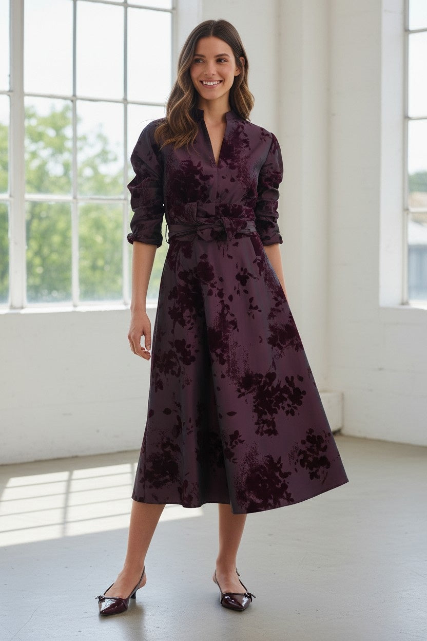 A woman stands in a bright room, smiling, with brown hair styled in loose waves. She wears the ROSSO35 Long Sleeve Taffeta Dress with Velvet by ROSSO 35 SILKY—a deep purple, floral-patterned midi dress crafted from printed flocked taffeta. Her outfit is completed with matching pointed shoes, making it perfect for a special occasion.