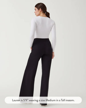 A woman stands against a plain background wearing the SPANX Perfect Pant Wide Leg in black—crafted from premium ponte fabric—paired with a white long-sleeve V-neck top and black heeled shoes.