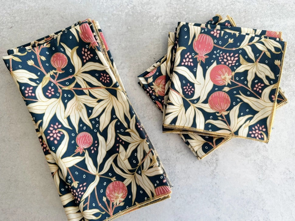 Two transparent plates, one yellow and one pink, rest on a light surface. Each displays DOT AND ARMY's Gooseberry Dinner Napkin—a dark blue cotton napkin with a beige, red, and green leaf pattern for stylish sustainable dining.