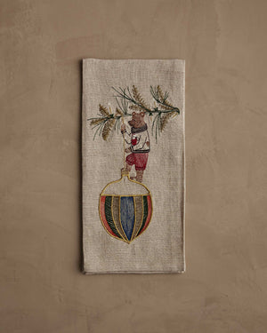 DECK THE HALLS BEAR LINEN TEA TOWEL featuring a bear in a sweater hanging a large, colorful, striped Christmas ornament from a pine branch on natural flax fiber.