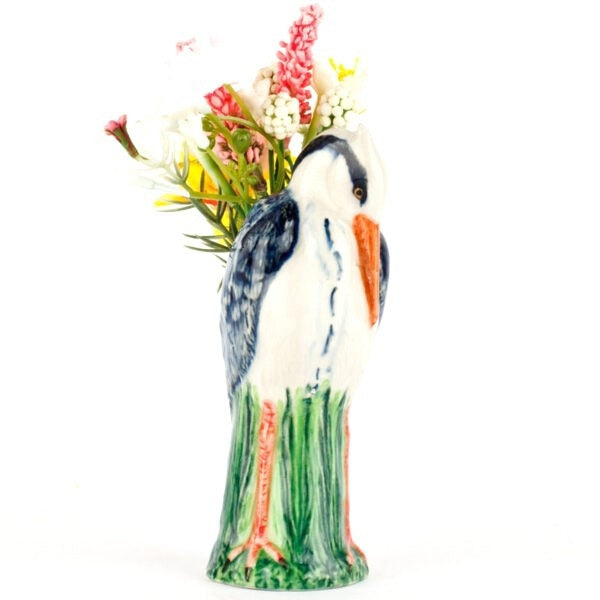 The QUAIL CERAMICS - HERON BUD VASE by QUAIL DESIGNS features a hand painted stoneware heron with blue and white feathers, orange beak, and green legs perched on a green base, shown against a plain white background.
