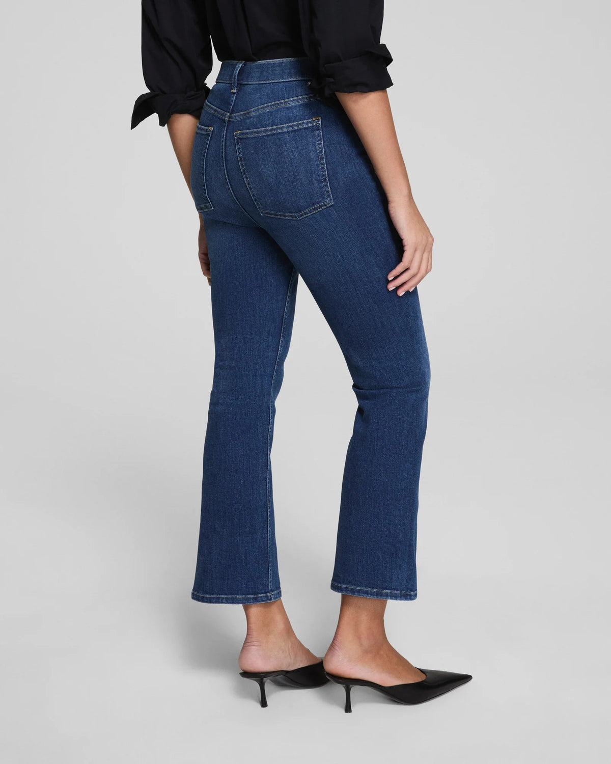 A woman with her hair pulled back stands against a plain background wearing SPANX Redefine Kick Flare Jeans in Medium Indigo, a black button-up shirt, black pointed-toe heels, and carries a black shoulder bag with one hand in her pocket.