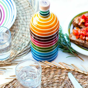 Introducing the MURAGLIA - EXTRA VIRGIN OLIVE OIL RAINBOW by ZIA PIA IMPORTS: a beautifully handcrafted ceramic jar with a cork stopper, featuring horizontal stripes in an array of vibrant colors like yellow, orange, red, green, blue, and pink. This decorative jar is ideal for storing Coratina extra virgin olive oil.