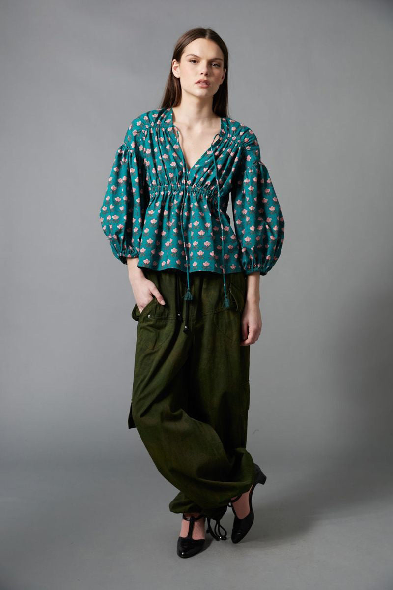 A person with long brown hair, seen from behind, wears the HOPE FOR FLOWERS Shirred Blouse and loose, dark green cargo pants with large pockets, standing against a plain gray background.