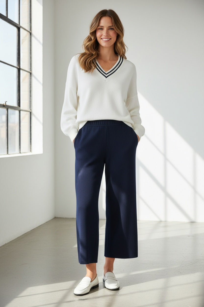 A woman with straight dark hair and a neutral expression stands against a white background, wearing a sleeveless white striped top, white sneakers, and the KINROSS CASHMERE Coverstitch Crop Cotton Blend Pant in light beige from KINROSS.