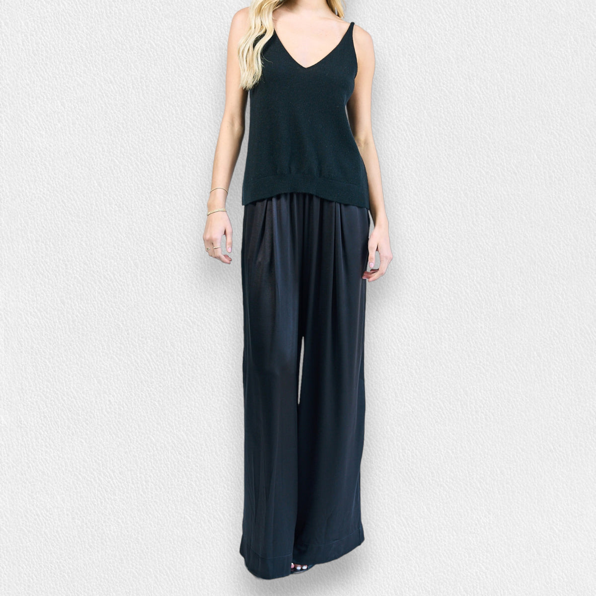A woman stands against a light textured background wearing ROSE &amp; CROWN’s SATIN TROUSER, paired with a sleeveless V-neck black top. Her effortlessly chic look is perfect for an evening out.