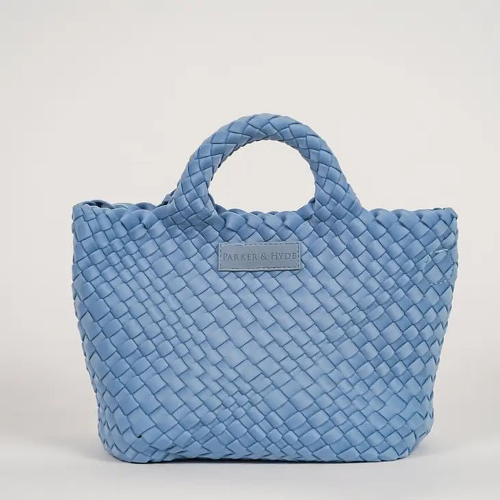 A woman with long brown hair, in a sleeveless cream knit top and blue jeans, holds a PARKER AND HYDE - MINI WOVEN TOTE BAG. The hand-woven tote by PARKER &amp; HYDE features a subtle checkered pattern with light blue accents.