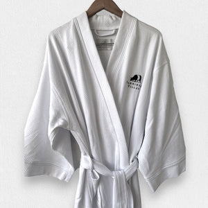 A neatly folded white kimono collar bathrobe adorned with a textured pattern. The bathrobe features an embroidered black bear and the text "Fearrington Village" on the bottom right side. Additionally, it boasts adjustable belt loops for a perfect fit. The label inside the collar reads "F".