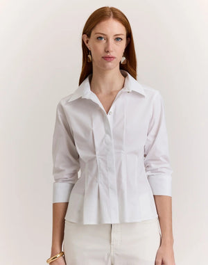 A woman with long red hair wears the HINSON WU - MARTINA 3/4 SLEEVE STRETCH POLISHED COTTON SHIRT IN WHITE, paired with white pants, gold earrings, and a gold bracelet, standing against a plain light background.