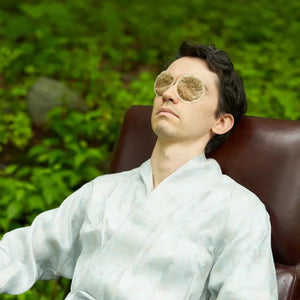 A person in a robe with a towel wrapped on their head relaxes on a green lounge chair, holding one BAUDELAIRE - EYES TEA BAG and wearing another over their other eye to help reduce dark circles and puffy eyes, surrounded by greenery.