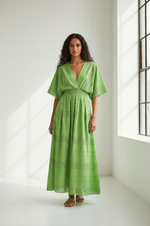 A person wears the MILVA SANGALLO DRESS, a light green cotton v-neck with eyelet details, short wide sleeves, and an ankle-length hem, standing against a plain white textured background.