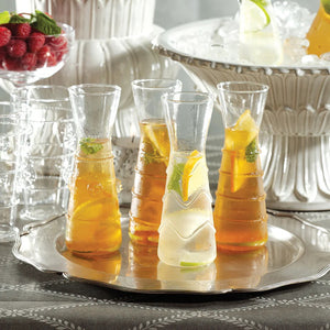 Six ZALLI SINGLE SERVE GLASS CARAFES by ZODAX, each featuring a unique etched design, are arranged on a reflective silver tray with a blurred green background.