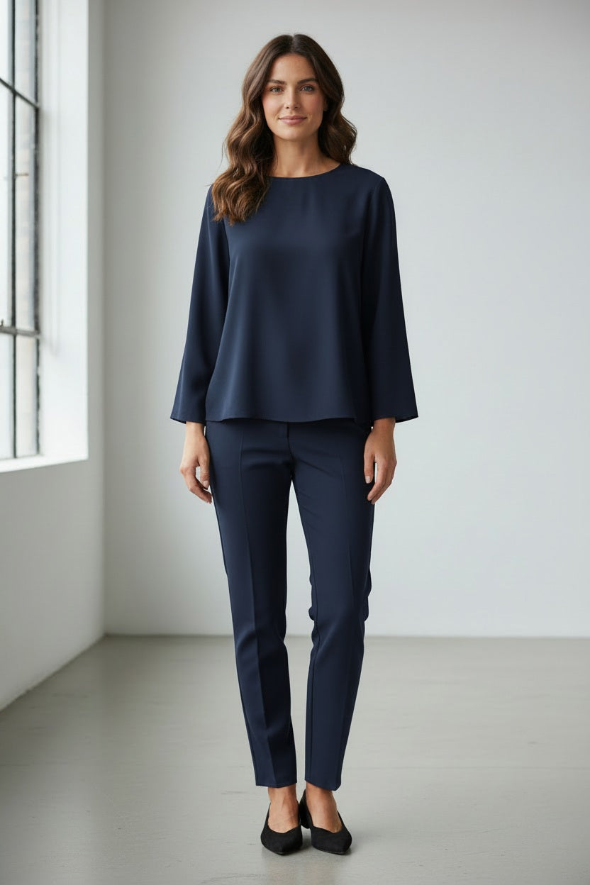 A person wears the MEIMEIJ TOP WITH KNOT BACK DETAIL in bright orange, paired with matching pants, standing against a plain light grey background. Their face is partially out of frame.