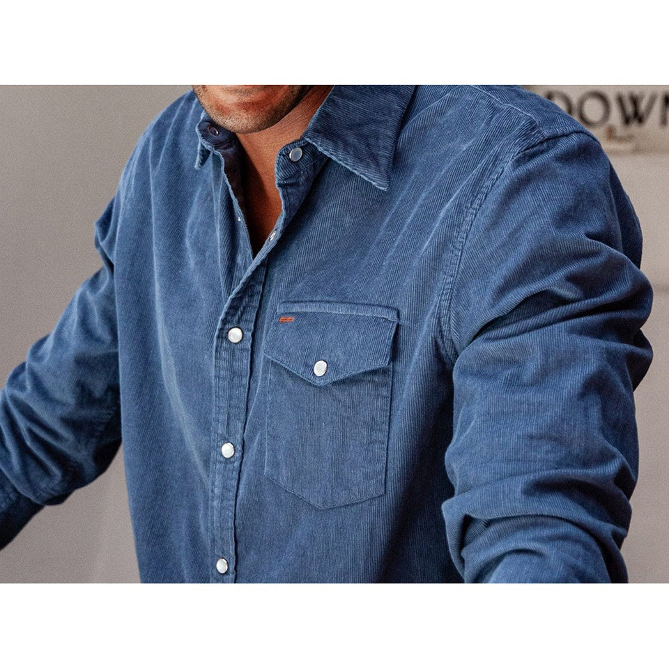 A man is wearing the CRIQUET Long Sleeve Corduroy Pearl Snap Shirt in Slate Blue by CRIQUET, featuring a chest pocket and rolled-up cuffs. He pairs it with brown pants against a white background.