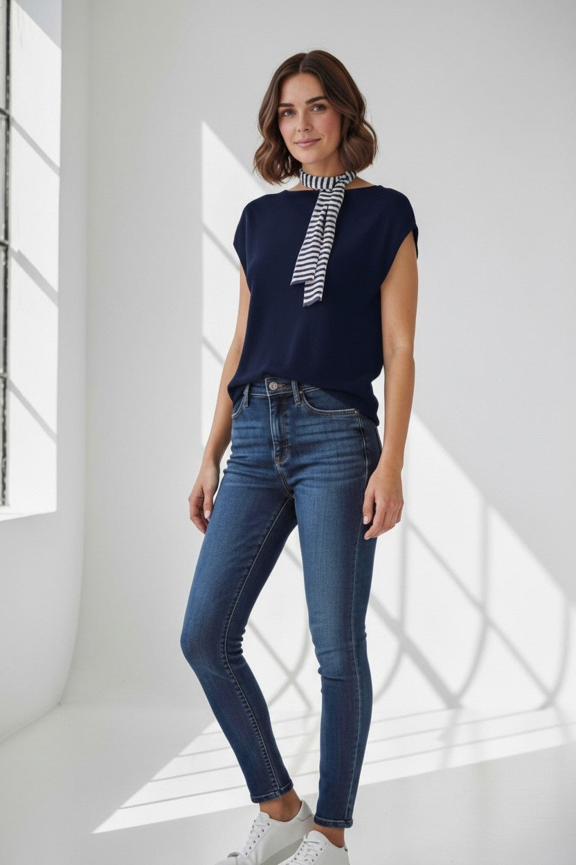 A woman is standing in a sunlit room near a large window with grid shadows on the floor, wearing the FLOOR Cotton Top with Stripe Neck Tie by FLOOR, paired with blue skinny jeans and white sneakers. She is smiling, showcasing the stylish black-and-white striped neck tie detail of her navy blouse.