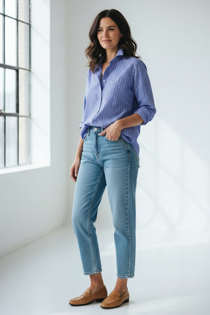 A woman stands in a sunlit room, smiling softly with wavy brown hair and one hand tucked into her pocket near a large window. She wears light blue jeans, tan loafers, and the FRANK & EILEEN EILEEN Relaxed Button Up Shirt in Italian Superluxe Cotton Multi Blue Stripe by FRANK & EILEEN. The shirt’s bust-flattering button placement adds a subtle touch of elegance to her effortlessly chic look.