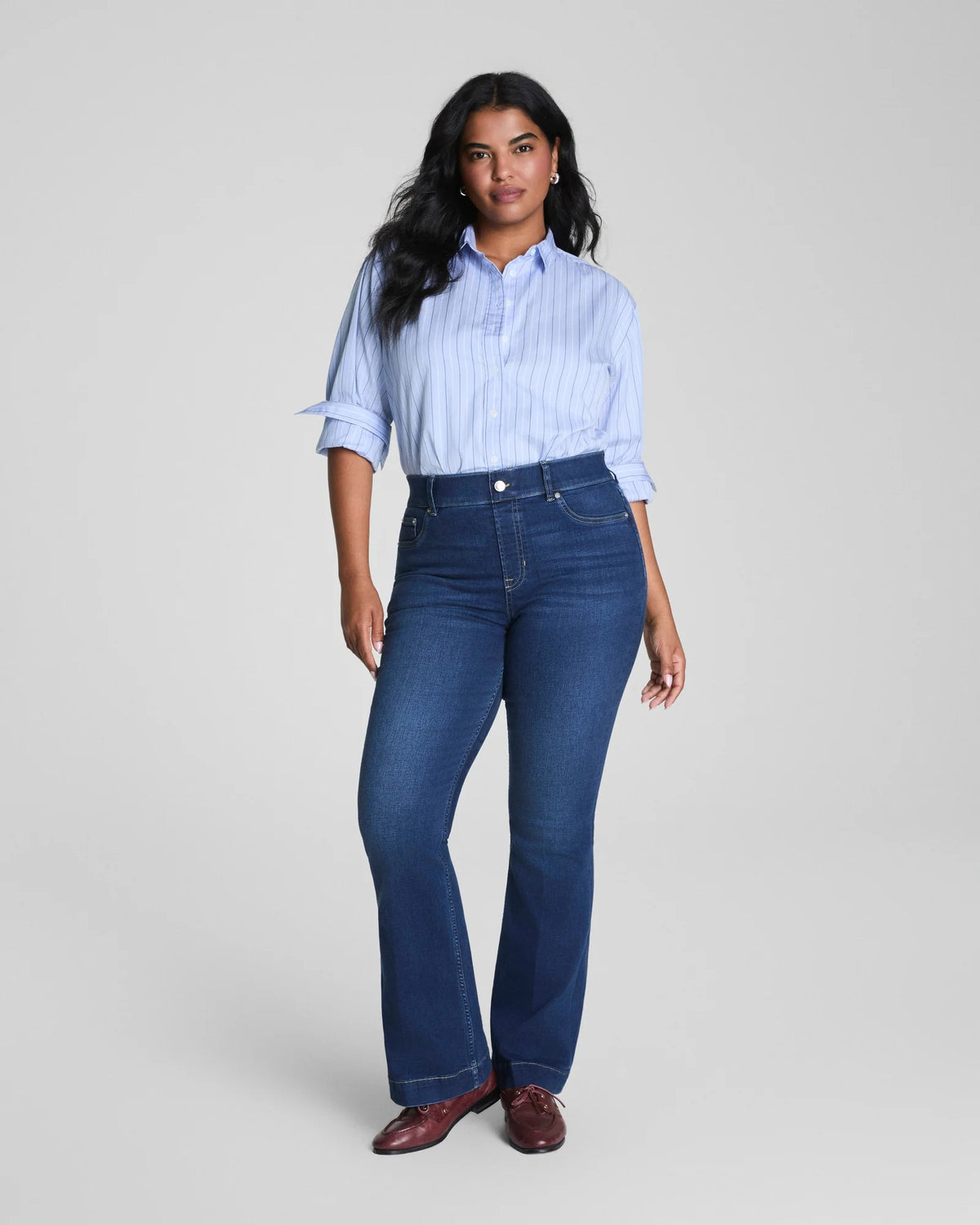 A woman poses against a plain light gray background wearing a blue striped button-up shirt with rolled sleeves, brown shoes, and the SPANX Shape Every Wear Pull On Flare Jean in Poseidon by SPANX, crafted from shaping denim.