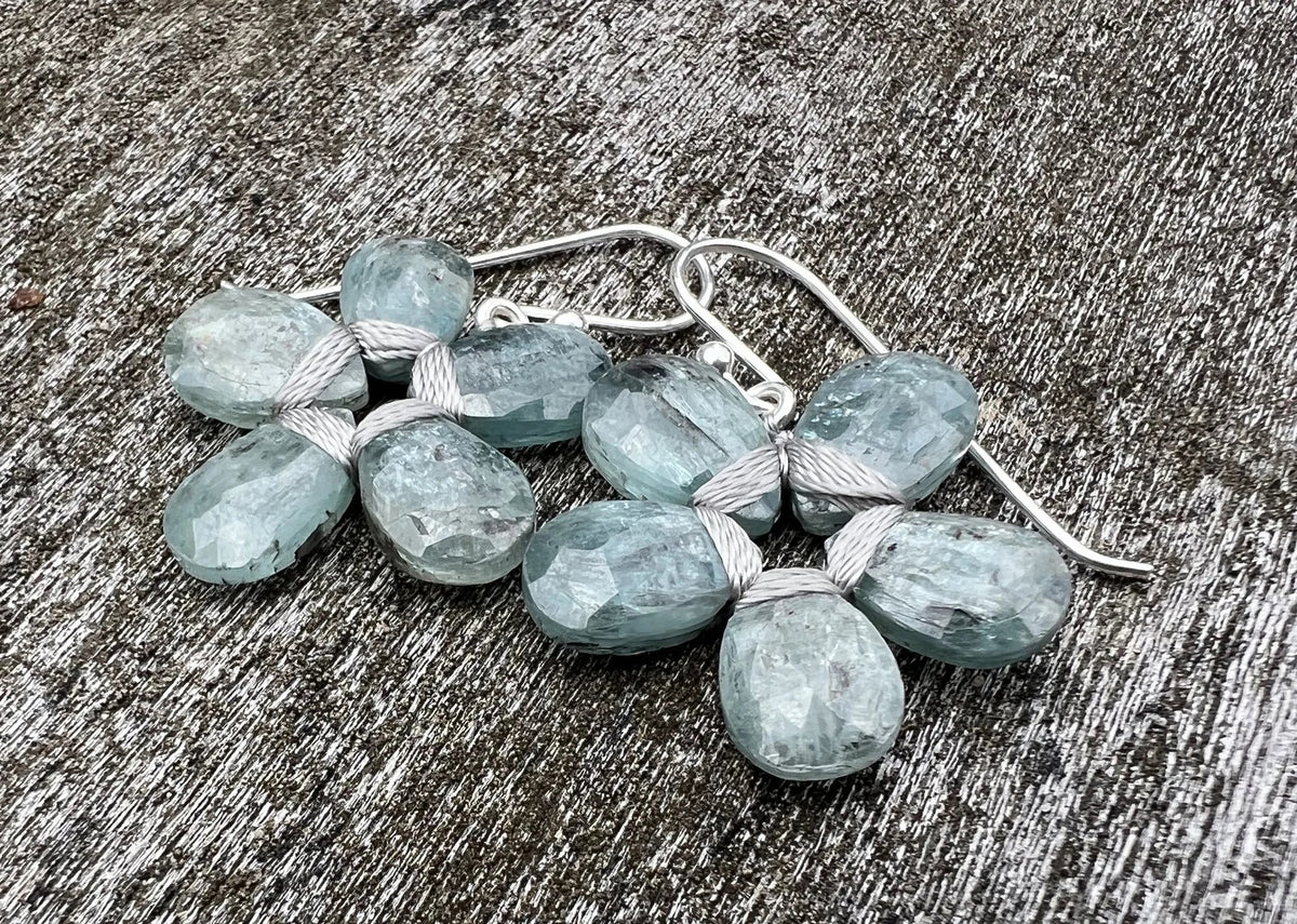 The ANN LIGHTFOOT - BLUE GREEN KYANITE FLOWER EARRINGS by ANN LIGHTFOOT, featuring faceted blue green kyanite clusters, are displayed on a weathered gray wooden surface.