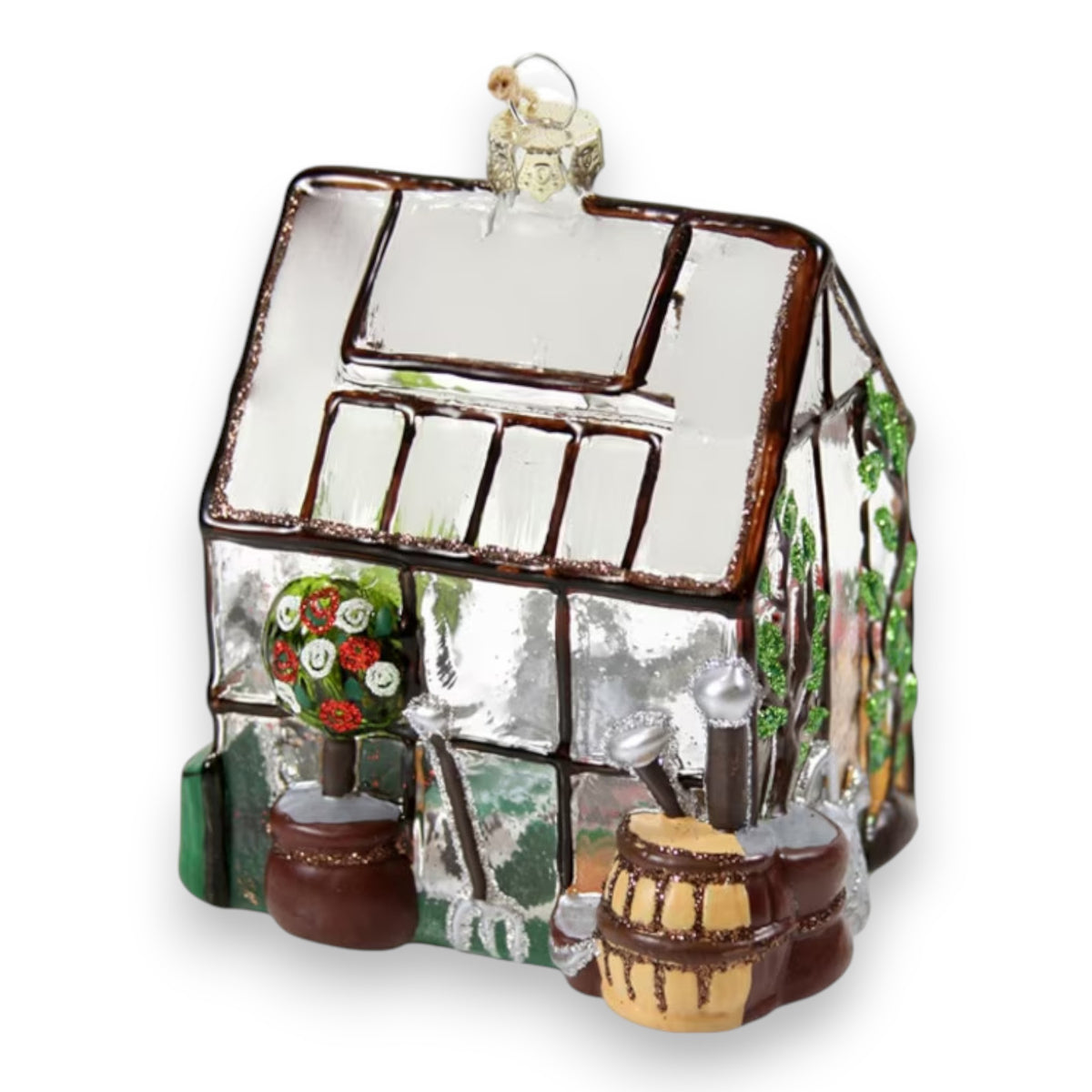 The CODY FOSTER GREENHOUSE ORNAMENT is a glass ornament featuring a greenhouse with brown trim, potted plants, barrels, a watering can, gardening tools, green vines, and a hanging loop at the top.