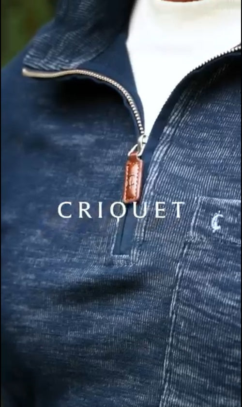 A man with short dark hair and a beard wears the CRIQUET - CROSSROADS Pullover in Moody Blue from CRIQUET over a white shirt and blue jeans. He stands facing forward against a plain white background.