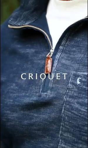 A man with short dark hair and a beard wears the CRIQUET - CROSSROADS Pullover in Moody Blue from CRIQUET over a white shirt and blue jeans. He stands facing forward against a plain white background.