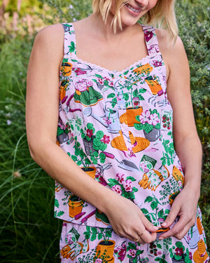 Wearing the PRINTFRESH Green Thumb Back to Bed Pajama Short Set—a vibrant, printed sleepwear ensemble from PRINTFRESH—a woman stands smiling in a lush, green garden, glancing to the side.