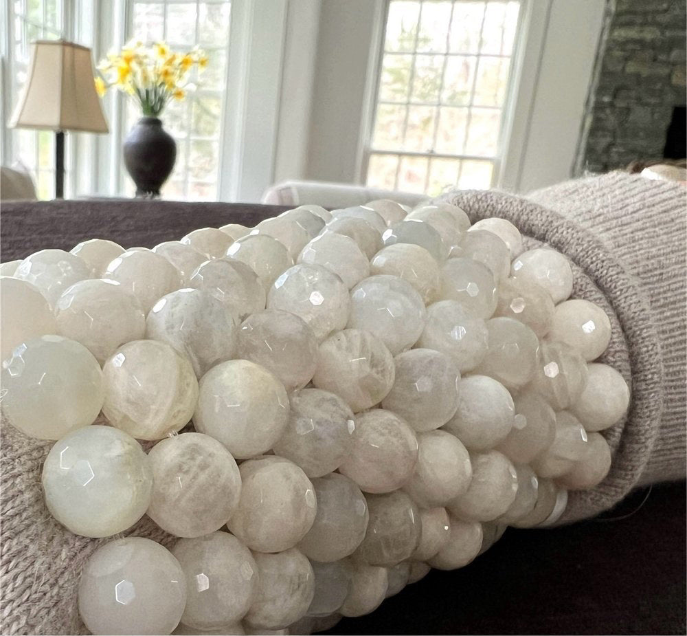 A detailed view of the ANN LIGHTFOOT Faceted 12mm White Moonstone Elastic Cuff Bracelet, showcasing a cluster of round, polished beads with a milky and translucent look. These beads give off a subtle sheen and are closely strung together on an elastic cuff, set against a plain white background.