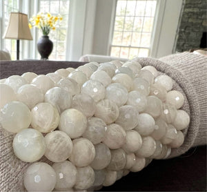 A detailed view of the ANN LIGHTFOOT Faceted 12mm White Moonstone Elastic Cuff Bracelet, showcasing a cluster of round, polished beads with a milky and translucent look. These beads give off a subtle sheen and are closely strung together on an elastic cuff, set against a plain white background.
