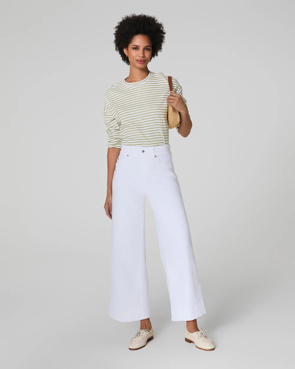 A person with short curly hair wears a striped long-sleeve shirt and high-waisted white SPANX PULL ON CROPPED WIDE LEG JEAN with TummyTamer Tech™. Against a plain background, they&#39;re holding a woven handbag and wearing beige shoes, exuding relaxed elegance.