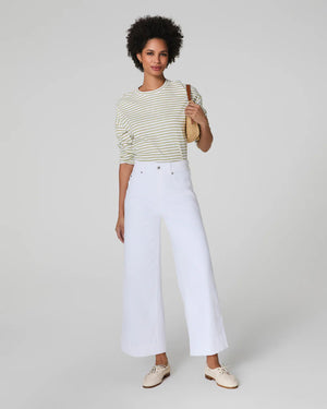 A person with short curly hair wears a striped long-sleeve shirt and high-waisted white SPANX PULL ON CROPPED WIDE LEG JEAN with TummyTamer Tech™. Against a plain background, they're holding a woven handbag and wearing beige shoes, exuding relaxed elegance.