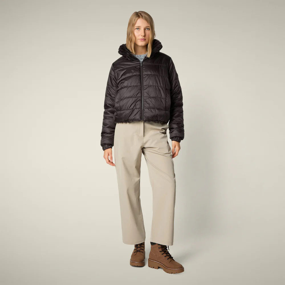 A woman models the SAVE THE DUCK - ELINA REVERSIBLE JACKET in beige fur, styled with a white skirt, orange tights, burgundy boots, and a matching clutch, standing against a plain light background.