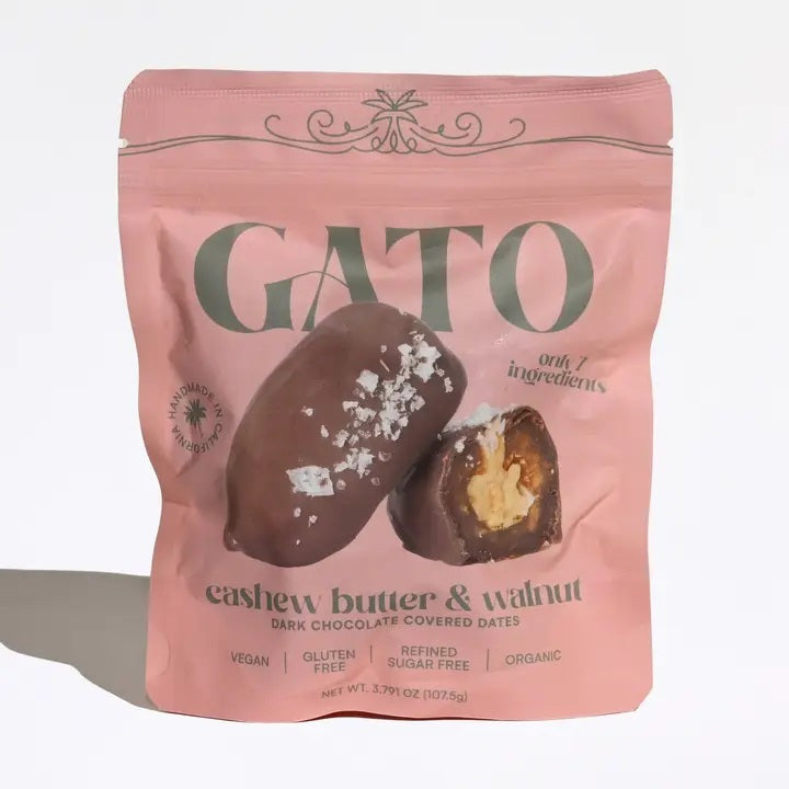A pink pouch of GATO DATES - CHOCOLATE COVERED CASHEW BUTTER & WALNUT DATES by GATO DATES, featuring an image of two chocolate pieces—one sliced open to reveal the filling. The packaging highlights that it’s vegan, gluten-free, organic, and proudly plant-based.