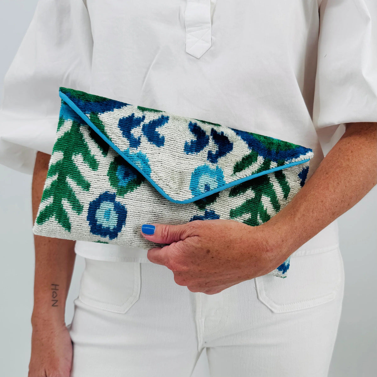 A person in a white outfit holds the BALDIZ - LILY CLUTCH IN AQUA VINE by BALDIZ, featuring blue, green, and cream designs. Their nails are painted blue and a small tattoo adorns their forearm.