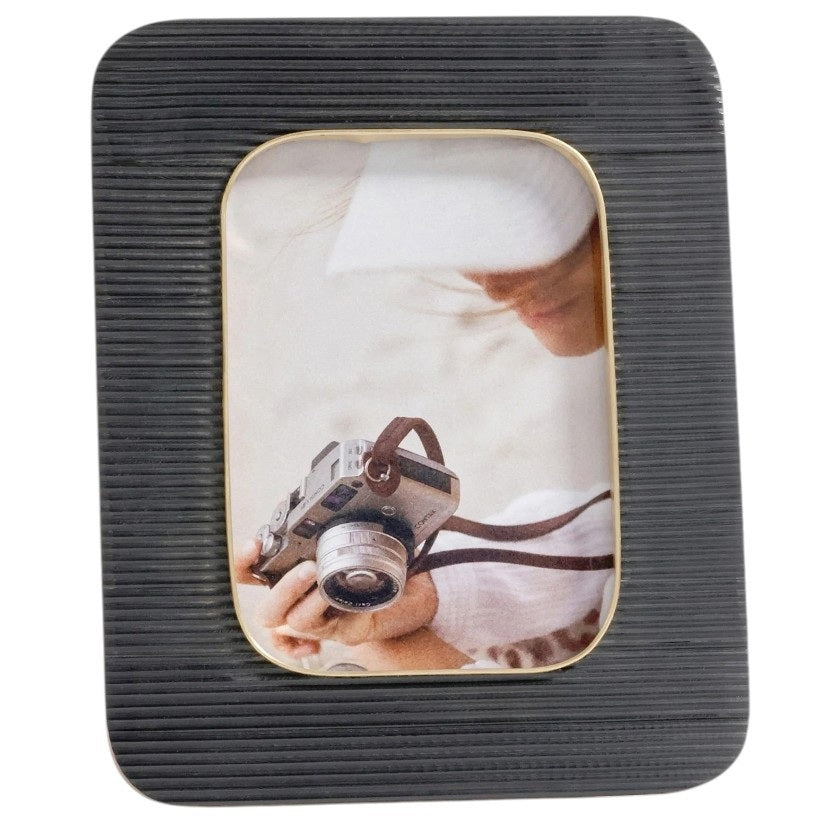 Two ZODAX BLACK RESIN RIBBED PHOTO FRAMES WITH BRASS ACCENT (5x7) display photos of a person in white, one with a camera. Each frame rests on beige boxes against a light blue background.