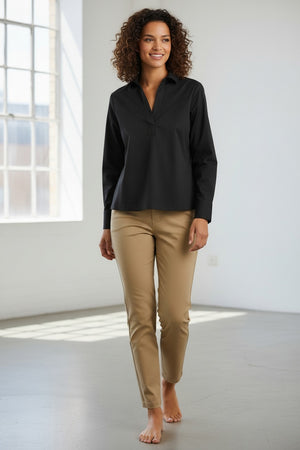 A woman stands barefoot indoors, smiling and looking to her left as natural light from a large window illuminates her effortlessly chic outfit. She wears the HINSON WU Monica Long Sleeve Popover with Buttons Down the Back, paired with tan stretch polished cotton pants, highlighting the relaxed fit of her ensemble.