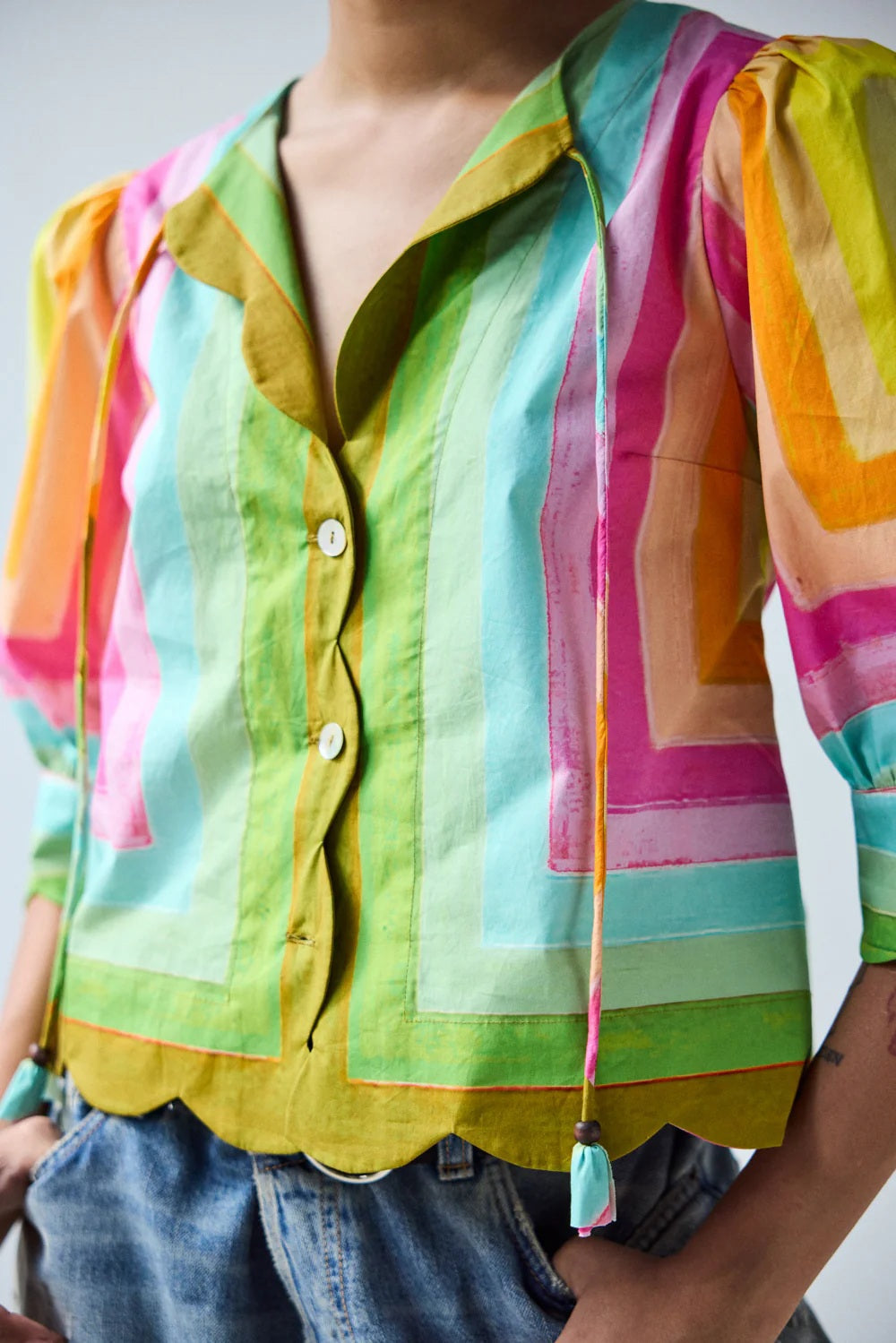 A person wears the HOPE FOR FLOWERS - SCALLOPED SHIRT IN CONCENTRIC SQUARES, featuring colorful geometric stripes and scalloped edges, paired with light blue jeans. The image highlights the torso.