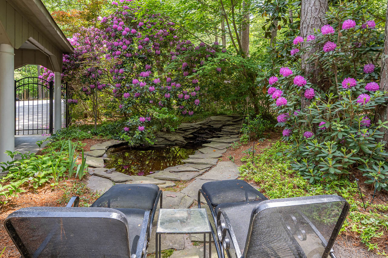 Two black mesh chairs and a small glass table face a stone path and small pond, surrounded by lush greenery and blooming purple rhododendrons in a shaded garden area.