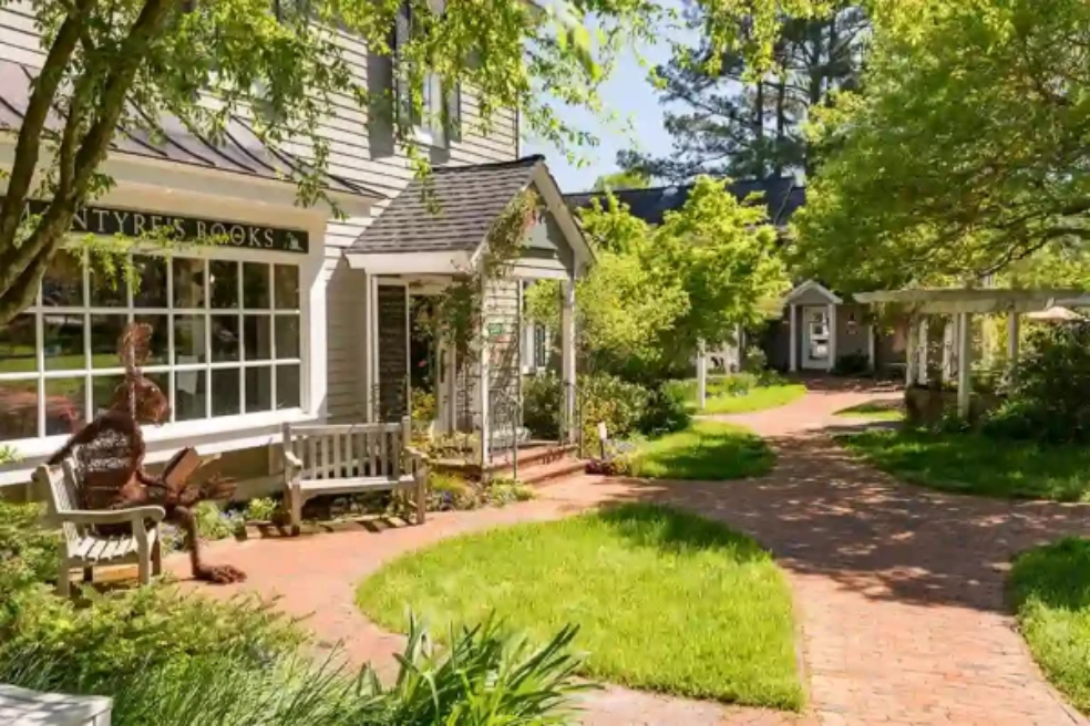 A quaint brick path winds through a lush garden, passing a bookstore with large windows and benches. Trees and greenery surround the area, creating a peaceful, inviting atmosphere.