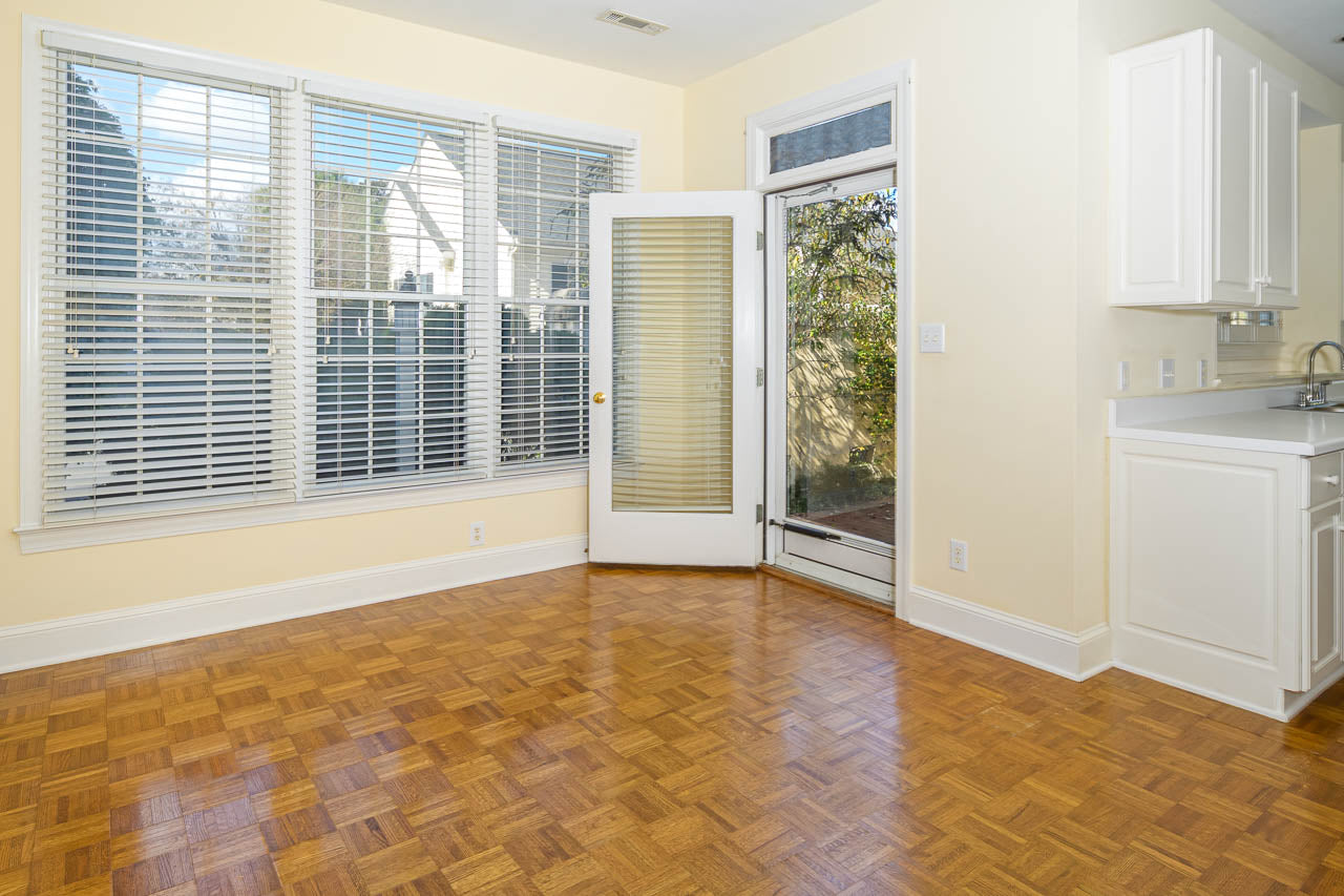 Sunlit room with large windows and white blinds, featuring a glass door leading outside. The space has light yellow walls, wood parquet flooring, and a partial view of a white kitchen with cabinets and a sink.