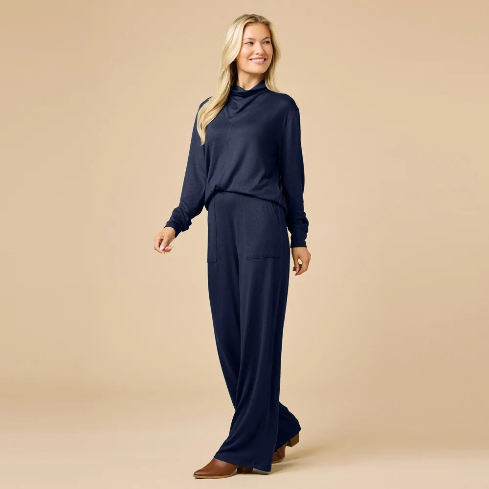 A woman wears a navy blue SOFTIES - DREAM JERSEY COWL NECK LOUNGE SET consisting of a long-sleeved top and wide-leg trousers.
