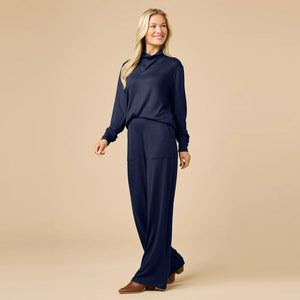A woman wears a navy blue SOFTIES - DREAM JERSEY COWL NECK LOUNGE SET consisting of a long-sleeved top and wide-leg trousers.