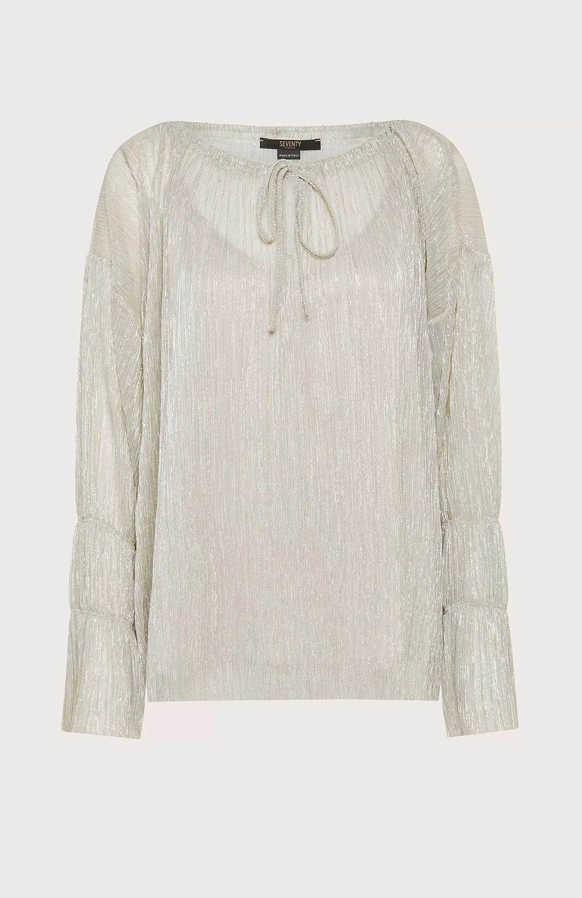 A person with shoulder-length hair stands against a light blue background, wearing the SEVENTY VENEZIA LUREX JERSEY BLOUSE by SEVENTY, which is a shimmery beige pleated blouse with long sleeves, paired with wide-leg beige trousers. The person gazes forward with a relaxed posture, one hand in their pocket.