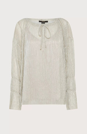 A person with shoulder-length hair stands against a light blue background, wearing the SEVENTY VENEZIA LUREX JERSEY BLOUSE by SEVENTY, which is a shimmery beige pleated blouse with long sleeves, paired with wide-leg beige trousers. The person gazes forward with a relaxed posture, one hand in their pocket.